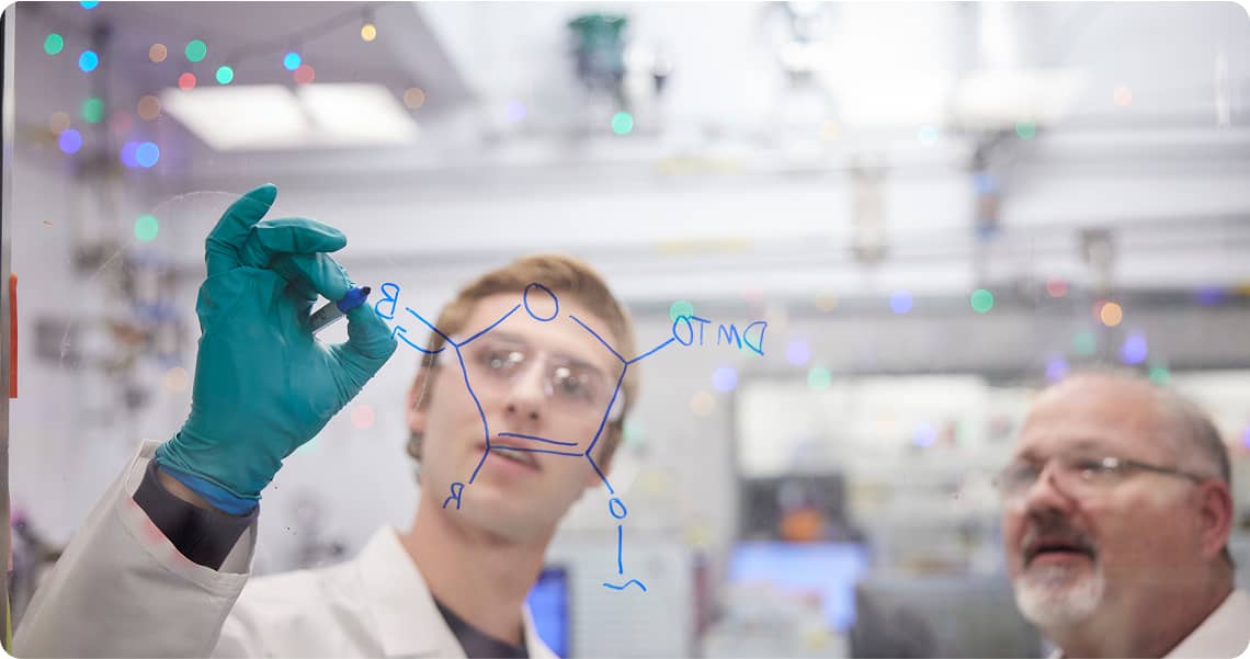 Two people in lab coats solving an equation on a clear, dry erase board