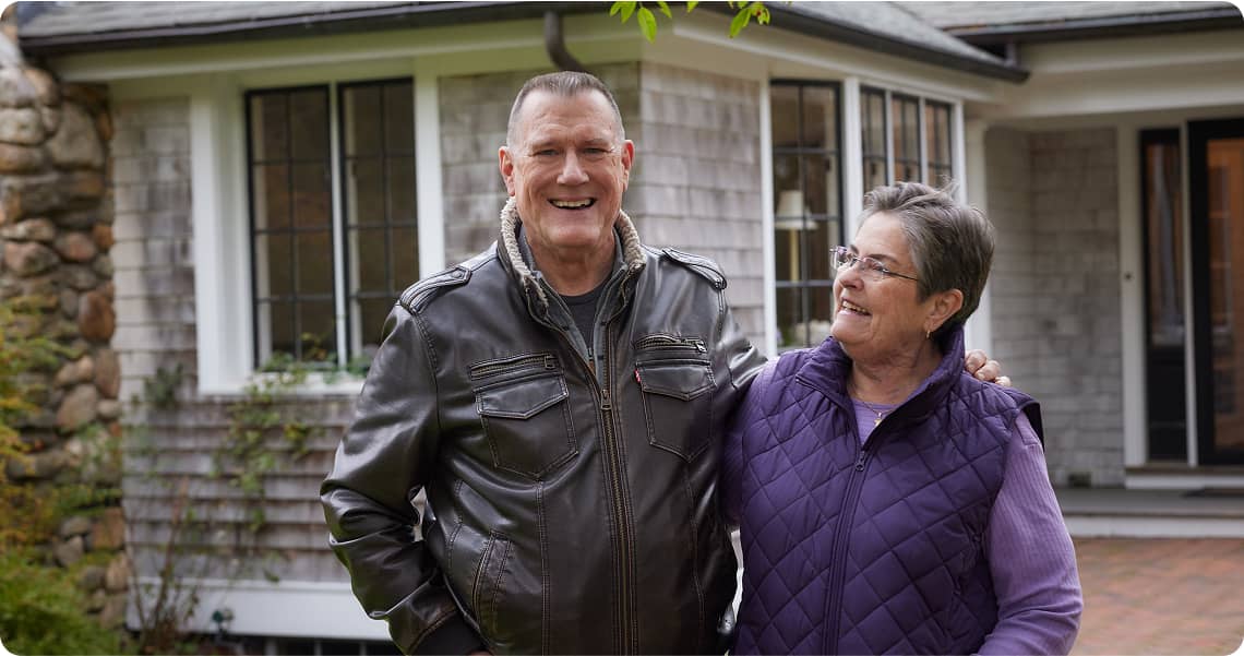 Mid shot of two people standing with an arm around each other smiling at each other and the camera
